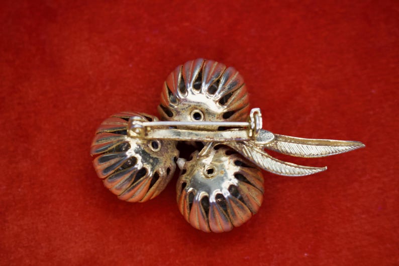 Puede incluir: Broche vintage con tres elementos esf&eacute;ricos plateados y texturizados. El broche tiene un cierre de alfiler y detalles decorativos en forma de hojas. El broche est&aacute; sobre un fondo rojo.