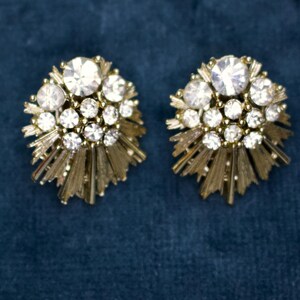 May include: A pair of vintage gold-toned clip-on earrings. Each earring features a cluster of clear, sparkling rhinestones surrounded by a sunburst design. The earrings are set against a dark blue background.