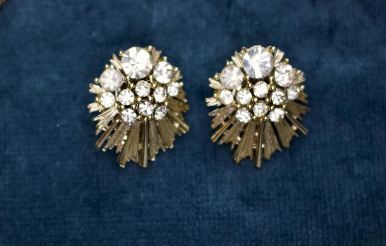 May include: A pair of vintage gold-toned clip-on earrings. Each earring features a cluster of clear, sparkling rhinestones surrounded by a sunburst design. The earrings are set against a dark blue background.