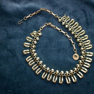 May include: A gold-toned necklace with a unique design, featuring elongated, teardrop-shaped elements. The necklace is set against a dark blue background, highlighting its metallic sheen. The clasp and chain are also gold, and a small pendant with a spiral design hangs near the clasp.