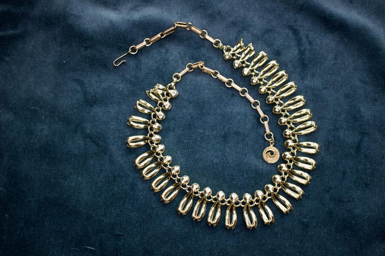 May include: A gold-toned necklace with a unique design, featuring elongated, teardrop-shaped elements. The necklace is set against a dark blue background, highlighting its metallic sheen. The clasp and chain are also gold, and a small pendant with a spiral design hangs near the clasp.