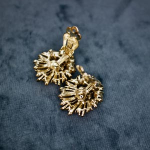 May include: A pair of gold-toned clip-on earrings. Each earring features a sunburst design with intricate detailing. The earrings are set against a dark teal background, highlighting their metallic finish and vintage style.