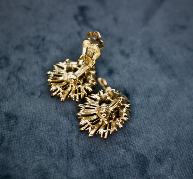 May include: A pair of gold-toned clip-on earrings. Each earring features a sunburst design with intricate detailing. The earrings are set against a dark teal background, highlighting their metallic finish and vintage style.