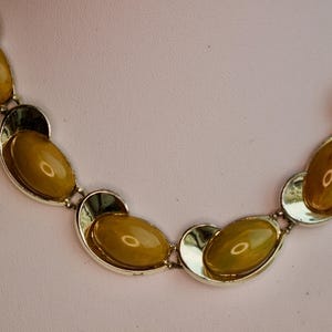 May include: A vintage necklace featuring oval, amber-colored stones set in silver-toned metal. The stones are linked together with a unique, open design. The necklace is displayed on a white surface against a soft pink background.
