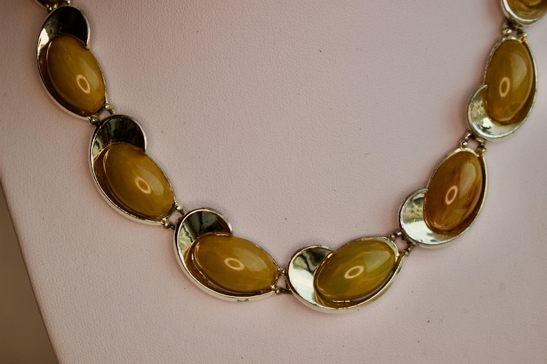 May include: A vintage necklace featuring oval, amber-colored stones set in silver-toned metal. The stones are linked together with a unique, open design. The necklace is displayed on a white surface against a soft pink background.