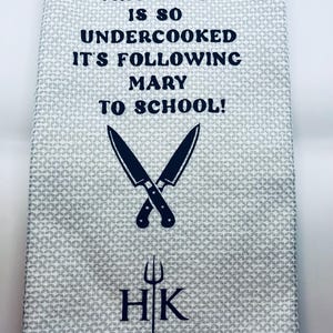 May include: A light gray kitchen towel with a repeating geometric pattern. The towel features the text "THIS LAMB IS SO UNDERCOOKED IT'S FOLLOWING MARY TO SCHOOL!" along with crossed knives, a fork, and the initials "HK".