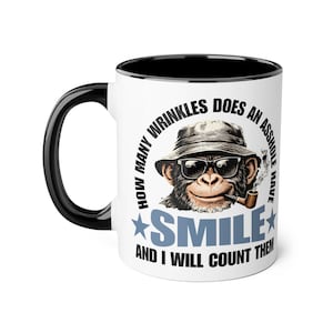 Puede incluir: Taza de cerámica blanca con asa y borde negros. La taza presenta un gráfico de un chimpancé con gafas de sol y sombrero, fumando en pipa. El texto en la taza dice "HOW MANY WRINKLES DOES AN ASSHOLE HAVE SMILE AND I WILL COUNT THEM."