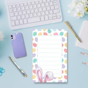 May include: Easter-themed notepad with a white lined interior, surrounded by colorful Easter eggs. The notepad features bunny ears at the bottom. A light blue desk holds a keyboard, phone, pen, and pencils.