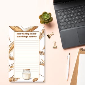 May include: A notepad with the text "just waiting on my sourdough starter" and lined pages. The notepad has a bread and wheat design. A small jar of starter is at the bottom. A laptop, pen, and binder clips are also on the pink surface.