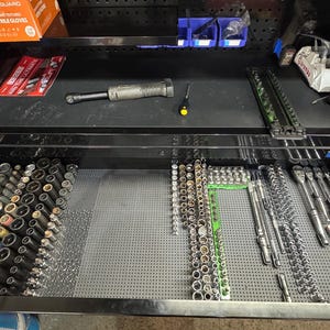 Może przedstawiać: Czarna szuflada skrzyni narzędziowej wypełniona różnymi narzędziami. Szuflada zawiera nasadki, grzechotki i inne narzędzia ręczne. Klucz dynamometryczny i śrubokręt znajdują się na górnej półce. Narzędzia są ułożone na szarej, perforowanej macie.