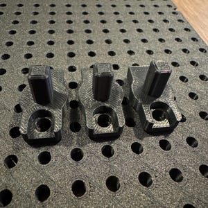 Peut inclure: Trois composants noirs imprimés en 3D, avec un dessus rectangulaire et une base incurvée, disposés sur une surface noire perforée. Les composants ont un trou au centre. L'arrière-plan est une surface sombre et texturée avec des trous circulaires espacés.