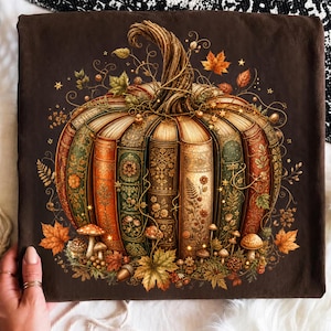 May include: A dark brown t-shirt featuring a graphic of a pumpkin constructed from vintage-style books with ornate gold and green spines. The design includes small mushrooms, scattered autumn leaves, and delicate golden vines surrounding the base.