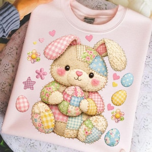 May include: A light pink sweatshirt featuring a patchwork bunny holding Easter eggs. The bunny has pink cheeks and ears, with floral and gingham patterns. Scattered around are hearts, flowers, and Easter eggs in various patterns.