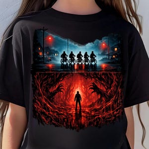 Puede incluir: Camiseta negra con un diseño dividido. La mitad superior muestra siluetas de ciclistas bajo un cielo nocturno. La mitad inferior muestra a una persona en una cueva roja con manos extendidas.