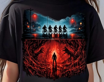 May include: Black t-shirt with a split design. The upper section shows silhouettes of cyclists under a night sky. The lower half depicts a person in a red cavern with reaching hands.