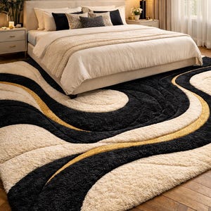May include: A large, plush area rug with a modern design. The rug features a wavy pattern in black, cream, and gold. The rug is placed in a bedroom setting, partially under a bed with neutral-toned bedding.
