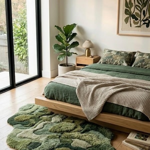 May include: A bedroom scene featuring a bed with green bedding and a beige waffle-knit blanket. A green, organic-shaped rug sits on the wooden floor. A framed botanical print hangs on the wall, and a potted plant is visible.