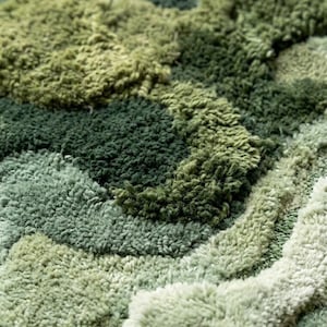 May include: Close-up of a textured rug with a pattern of varying shades of green. The rug's surface is made of soft, looped fibers, creating a plush, tactile appearance. The design features a layered, organic motif, resembling a landscape.