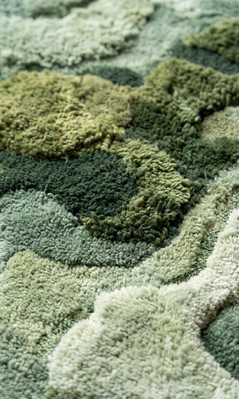 May include: Close-up of a textured rug with a pattern of varying shades of green. The rug's surface is made of soft, looped fibers, creating a plush, tactile appearance. The design features a layered, organic motif, resembling a landscape.