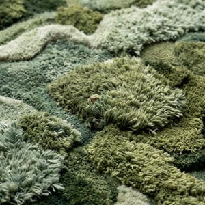 May include: A close-up view of a plush, textured rug in various shades of green. The rug's design features a layered, organic pattern with varying pile heights, creating a three-dimensional effect. The color palette ranges from light sage to deep forest green.