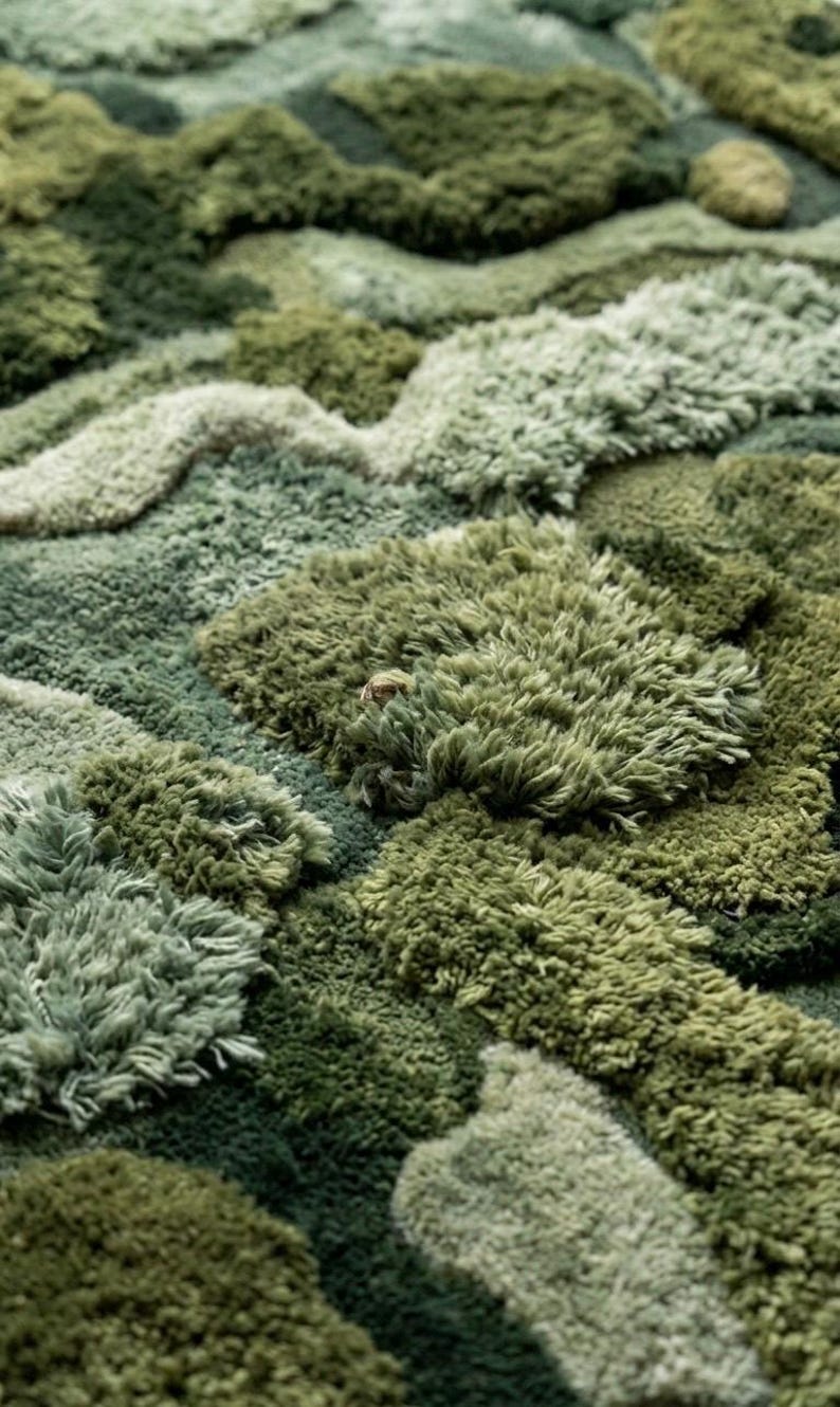 May include: A close-up view of a plush, textured rug in various shades of green. The rug's design features a layered, organic pattern with varying pile heights, creating a three-dimensional effect. The color palette ranges from light sage to deep forest green.