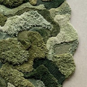 May include: A textured, abstract rug in shades of green, resembling a landscape. The rug features varying pile heights and textures, creating a dimensional effect. The design incorporates multiple shades of green, from light to dark, with a unique, organic shape.