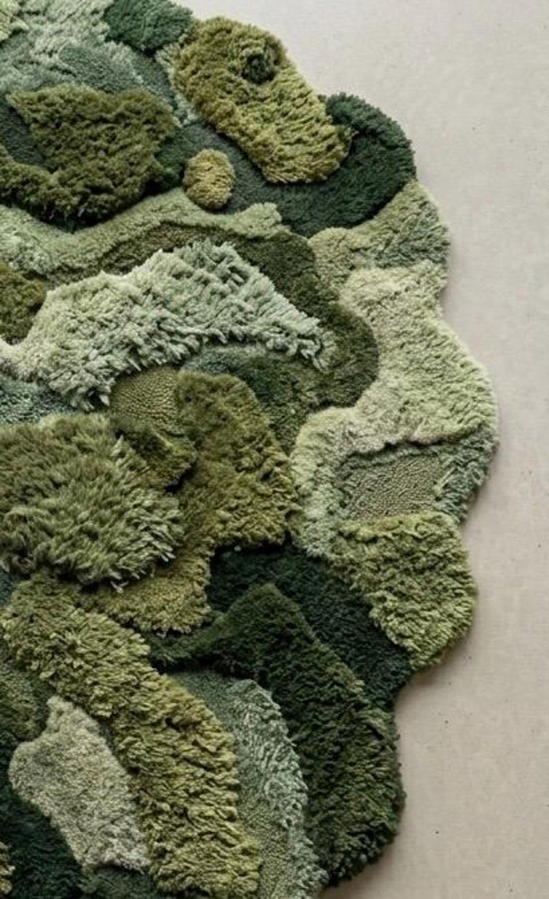 May include: A textured, abstract rug in shades of green, resembling a landscape. The rug features varying pile heights and textures, creating a dimensional effect. The design incorporates multiple shades of green, from light to dark, with a unique, organic shape.