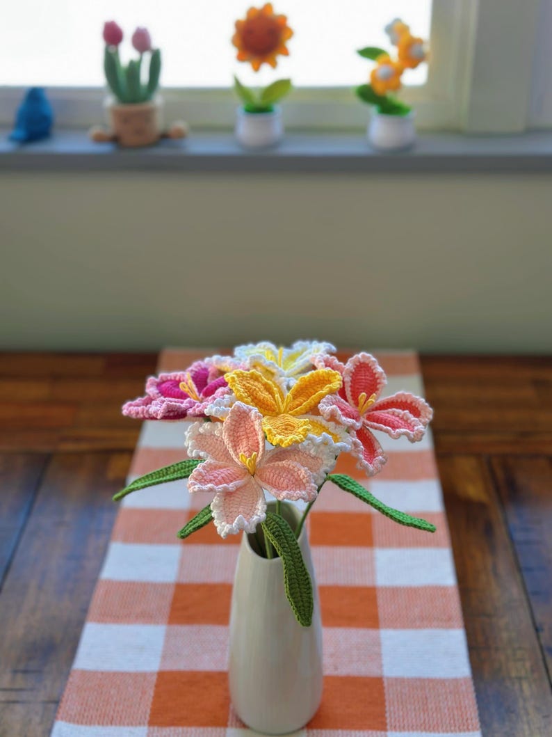 Puede incluir: Un ramo de flores de ganchillo hechas a mano en un jarr&oacute;n blanco, colocado sobre un camino de mesa con un patr&oacute;n de cuadros naranja y blanco. Las flores son en tonos de rosa, amarillo y naranja con tallos y hojas verdes. Flores de ganchillo decorativas son visibles en el fondo.