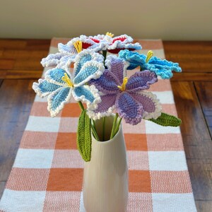 Puede incluir: Un ramo de flores de ganchillo hechas a mano en un jarr&oacute;n blanco. Las flores son en tonos de azul, morado y rojo con centros amarillos y bordes blancos. El jarr&oacute;n est&aacute; sobre un camino de mesa a cuadros naranja y blanco.