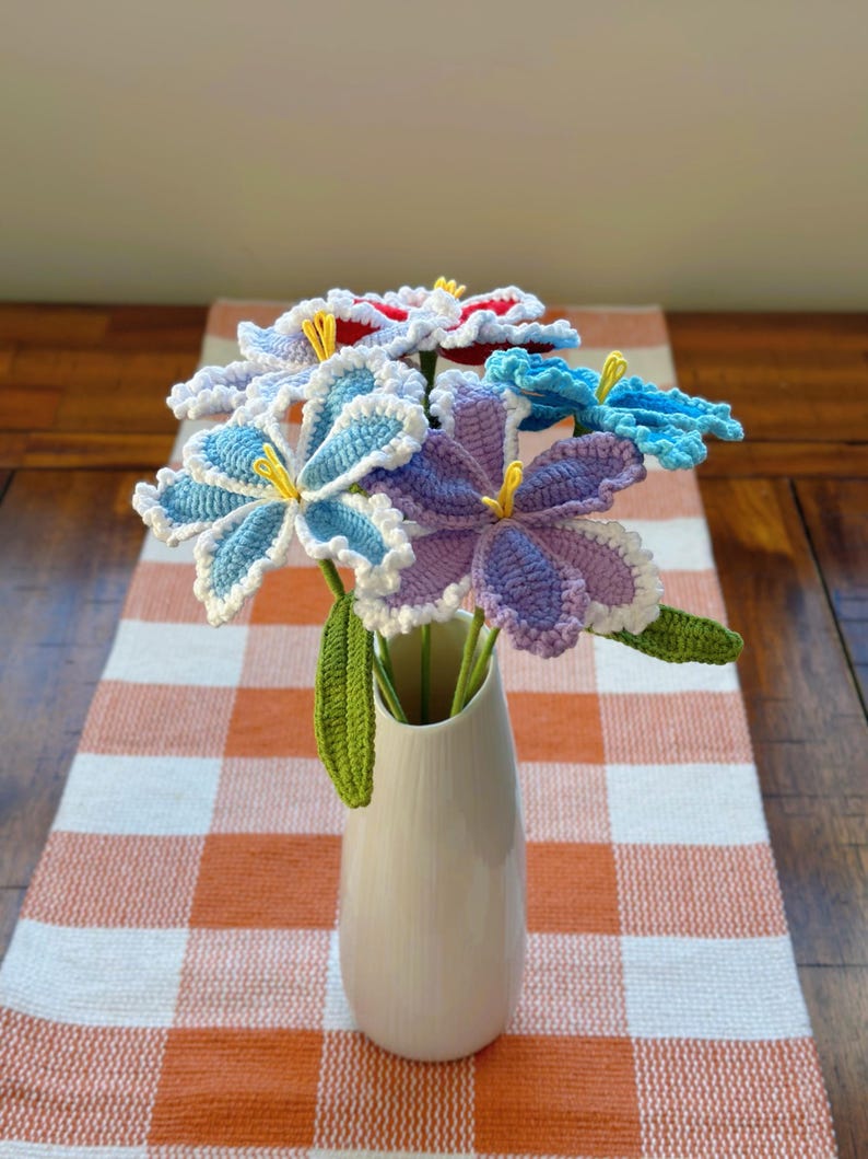 Puede incluir: Un ramo de flores de ganchillo hechas a mano en un jarr&oacute;n blanco. Las flores son en tonos de azul, morado y rojo con centros amarillos y bordes blancos. El jarr&oacute;n est&aacute; sobre un camino de mesa a cuadros naranja y blanco.