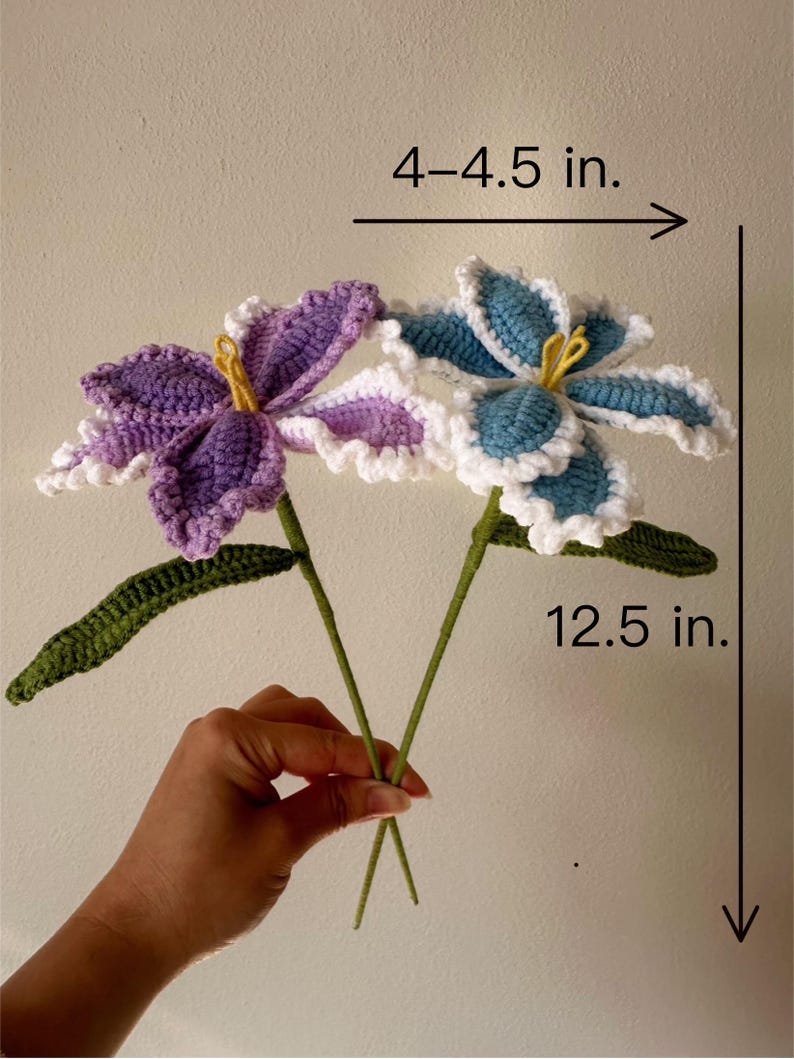 Puede incluir: Dos flores de ganchillo, una morada y otra azul, con detalles blancos y amarillos. Las flores miden aproximadamente 10-11 cm de ancho y 32 cm de alto. Los tallos y hojas verdes completan el arreglo floral hecho a mano.