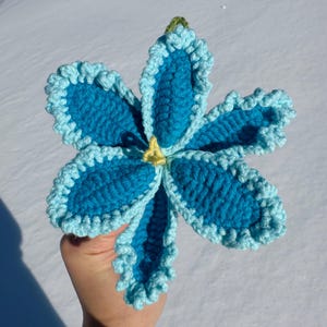 Puede incluir: Una flor hecha a mano, de ganchillo, en tonos azules con un borde rizado azul claro. El centro es amarillo y el tallo es verde. La flor es un art&iacute;culo &uacute;nico, hecho a mano.
