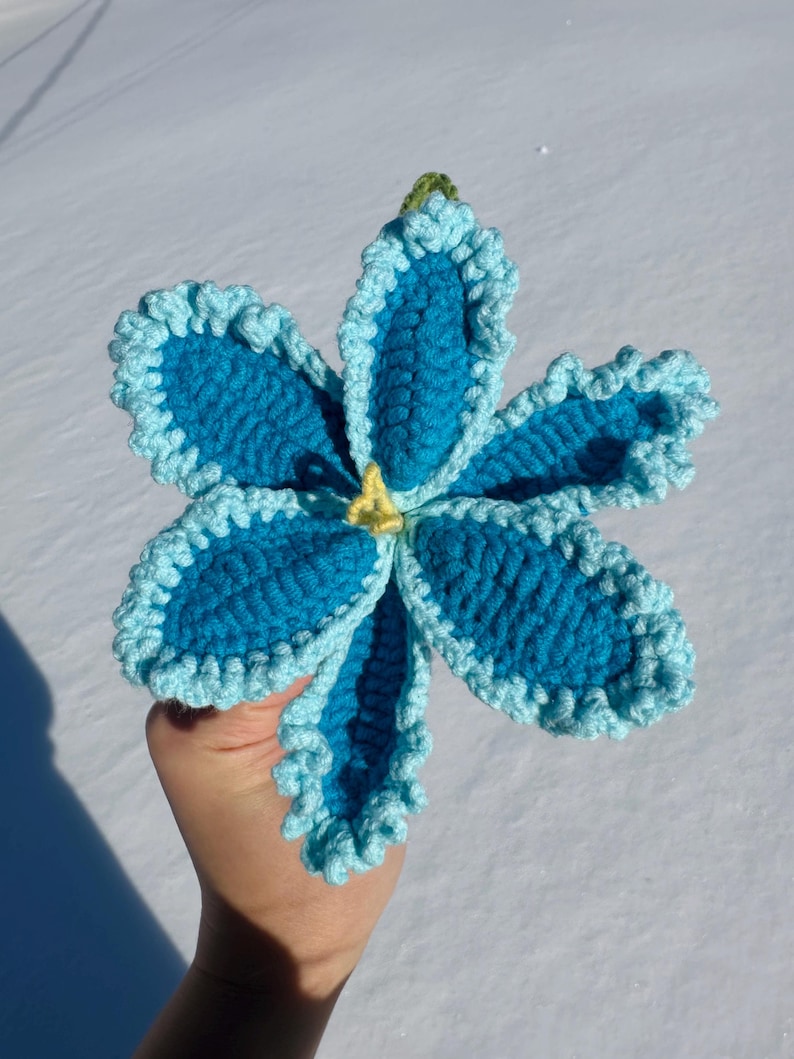 Puede incluir: Una flor hecha a mano, de ganchillo, en tonos azules con un borde rizado azul claro. El centro es amarillo y el tallo es verde. La flor es un art&iacute;culo &uacute;nico, hecho a mano.