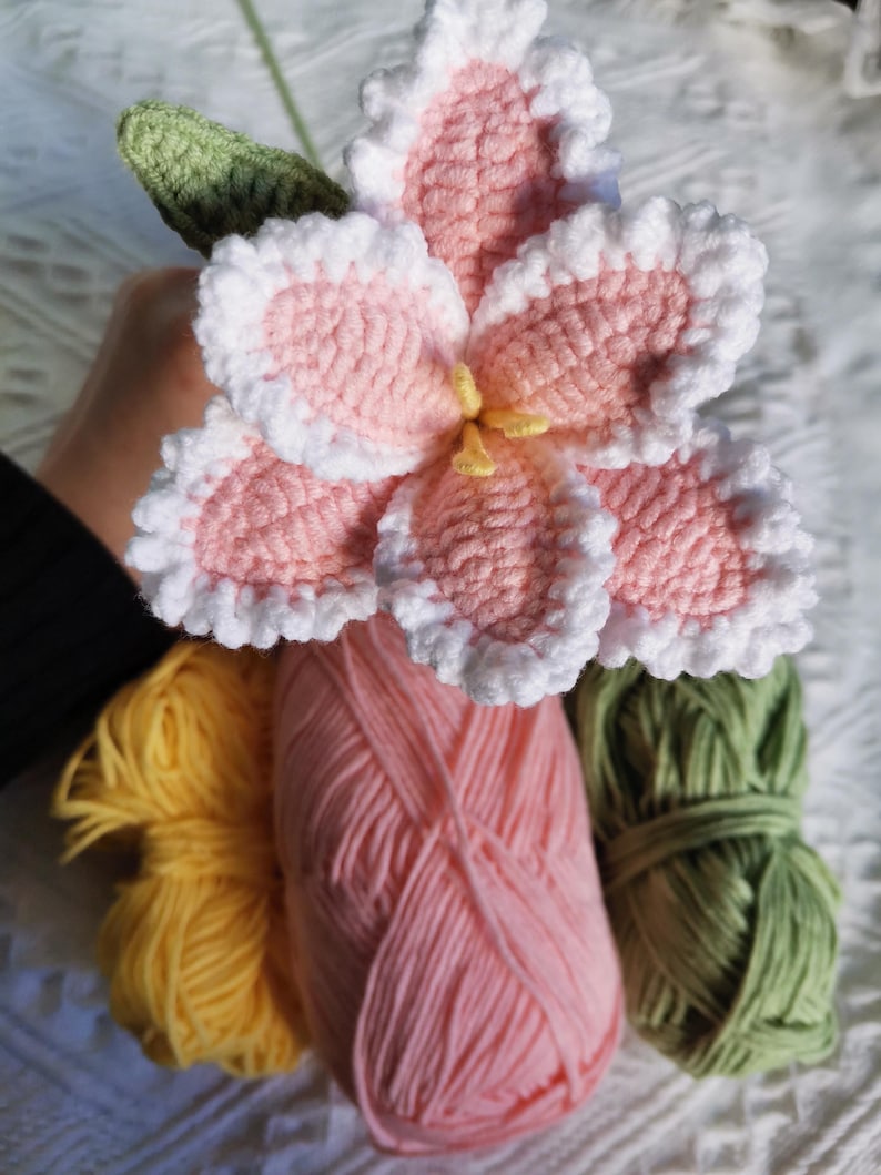 Puede incluir: Una flor de ganchillo hecha a mano con p&eacute;talos rosas, borde blanco y centro amarillo. Acompa&ntilde;ada de ovillos de lana amarilla, rosa y verde, lo que sugiere un proyecto de manualidades. La flor tiene una hoja verde.