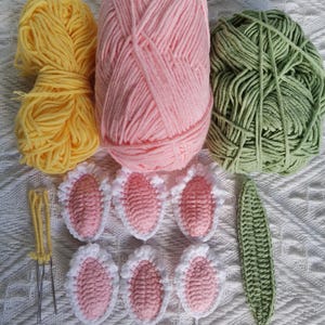Puede incluir: Una colecci&oacute;n de ovillos de lana en amarillo, rosa y verde, junto con formas de orejas de conejo y una hoja de ganchillo. La lana est&aacute; dispuesta con agujas de ganchillo, y las orejas de conejo son rosas con ribete blanco. La hoja es verde.