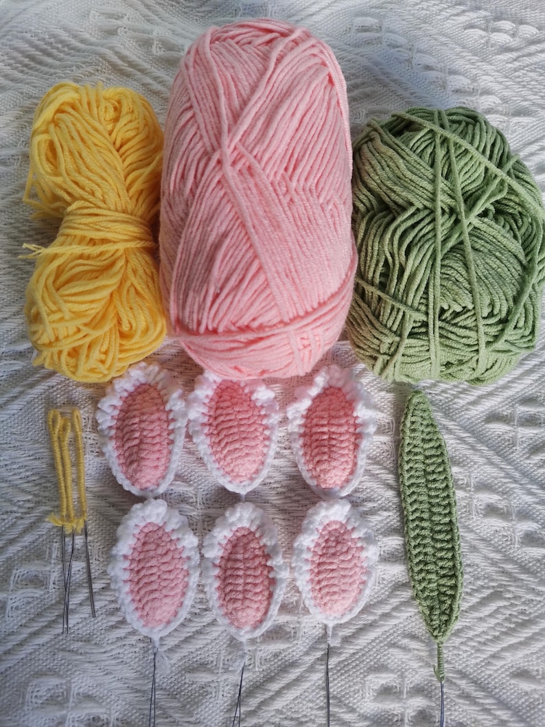Puede incluir: Una colecci&oacute;n de ovillos de lana en amarillo, rosa y verde, junto con formas de orejas de conejo y una hoja de ganchillo. La lana est&aacute; dispuesta con agujas de ganchillo, y las orejas de conejo son rosas con ribete blanco. La hoja es verde.