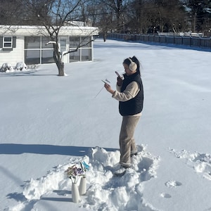 Puede incluir: Dos jarrones de cer&aacute;mica blanca con flores artificiales de colores est&aacute;n en la nieve. Una persona est&aacute; de pie en la nieve, sosteniendo un tel&eacute;fono y un objeto peque&ntilde;o. La escena es al aire libre en un d&iacute;a soleado con una casa al fondo.