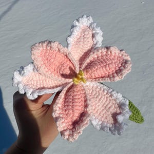 Puede incluir: Una flor de ganchillo hecha a mano en tonos rosa y blanco, con un centro amarillo y una hoja verde. La flor tiene bordes con volantes y se sostiene sobre un fondo nevado.