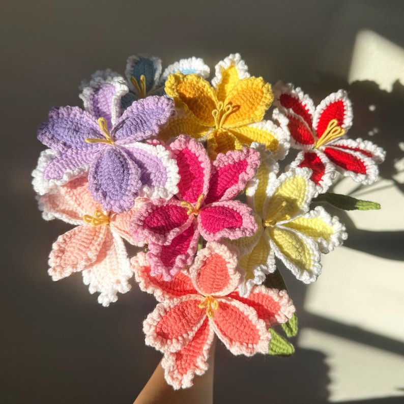 Puede incluir: Un ramo de flores de ganchillo hechas a mano en varios colores, incluyendo morado, rosa, amarillo y rojo. Cada flor tiene un dise&ntilde;o detallado de p&eacute;talos con bordes blancos y estambres amarillos. Las flores est&aacute;n unidas, creando una exhibici&oacute;n vibrante y colorida.
