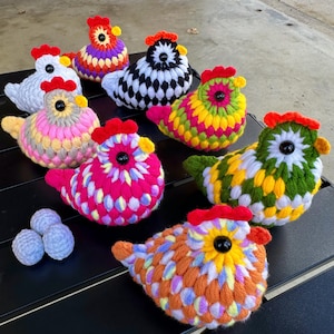 Puede incluir: Una colección de gallinas de ganchillo hechas a mano en varios colores, incluyendo blanco, rosa, amarillo, verde y naranja. Cada gallina tiene una cresta roja y un ojo negro. También se ven tres pequeños huevos de ganchillo azul claro.