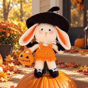 Puede incluir: Una muñeca de conejo de ganchillo blanca que lleva un sombrero de bruja negro y un vestido naranja. El conejo tiene orejas largas con secciones interiores naranjas, mejillas rosadas y sostiene un pequeño cubo de calabaza con una cara negra.