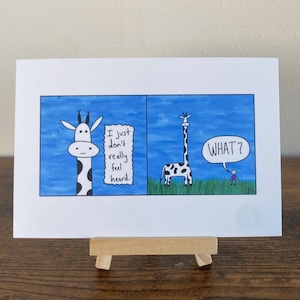 May include: A framed art print with a two-panel cartoon about giraffes. The first panel shows a giraffe with the text "I just don't really feel heard." The second panel shows a giraffe and a person with the text "WHAT?" The background is blue.