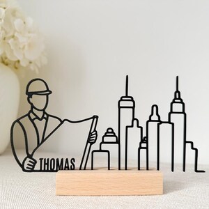 Op de afbeelding: Zwarte metalen bureau-decoratie met een architectfiguur met een blauwdruk en een skyline-silhouet. De naam "THOMAS" is zichtbaar. De basis is van lichtgekleurd hout. Een decoratief item.