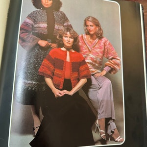 May include: Three women wearing different knitted sweaters. The woman in the center is wearing a red and brown striped sweater with a black skirt. The woman on the left is wearing a multi-colored striped sweater with a black skirt. The woman on the right is wearing a pink and white striped sweater with a gray skirt.
