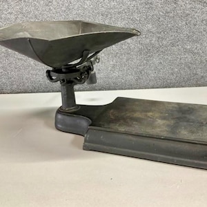 Puede incluir: Una antigua báscula de metal gris oscuro con una gran bandeja cuadrada y una base rectangular. La bandeja está suspendida sobre la base por una serie de soportes curvos y ornamentados. La báscula parece desgastada, con un aspecto ligeramente envejecido.