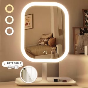 May include: A rectangular vanity mirror with a white frame and integrated LED lighting. The mirror features a touch-sensitive power button on the glass surface and sits on a white base tray. A small circular inset shows a data cable for charging.