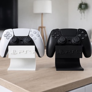 Könnte beinhalten: Zwei PlayStation 5-Controller, einer weiß und einer schwarz, auf passenden Ständern. Die Ständer zeigen das PS5-Logo. Die Controller befinden sich auf einer hellen Holzoberfläche.