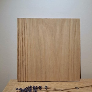 May include: A rectangular wooden board with a light brown grain pattern, standing upright on a wooden surface. A few sprigs of dried lavender are placed in front of the board, adding a touch of color and texture. The background is a plain white wall.