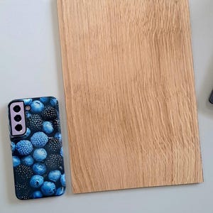 May include: A smartphone case with a blue and black berry pattern rests beside a rectangular piece of light brown wood. The wood grain is visible. The phone case has a camera module with three lenses.