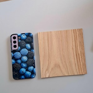 May include: A smartphone case with a vibrant blue and black berry design, alongside a small, square piece of light-colored wood. The case features a camera array. The wood has a natural grain pattern.